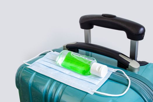 A green luggage with a face mask and a hand sanitizer on a white background. Concept traveling during pandemic. Travel covid 19.
