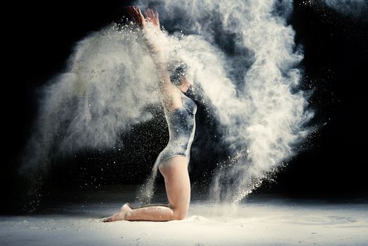 Caucasian woman with an athletic body sits on the floor and throws white flour up, dust flies in different directions, athlete is dressed in a black bodysuit