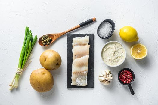 Traditional fish and chips ingredients recipe raw cod fillets on stone slate batter, potatoe, tartar sauce, lemon, capers , green herbs garlic, salt, peppercorns on white stone background flat lay top view .