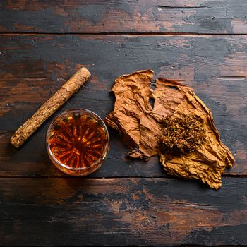 Cut tobacco and tobacco leaves with cigar and whiskey rum on wood background on vintage dark table. overhead shot top view.