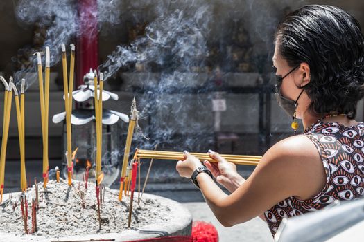 Asian woman wearing a medical mask to protect from infection of virus. Is burning incense sticks in a Chinese temple. Mask to protect herself from coronavirus is seen at the city. Selective focus.
