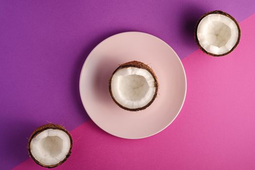 Coconut half in pink plate with nut fruits on violet and purple plain background, abstract food tropical concept, top view