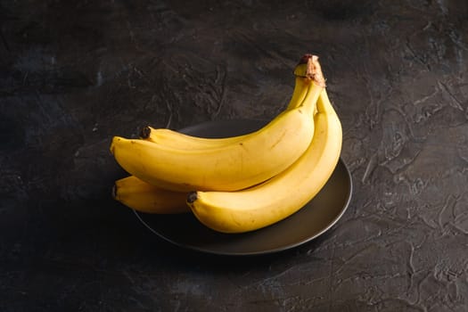 Banana fruits in black plate on dark textured background, angle view