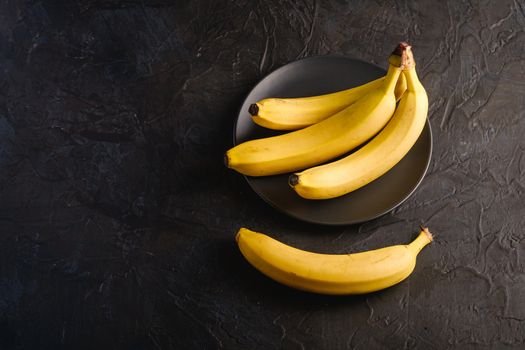 Banana fruits in black plate on dark textured background, angle view copy space