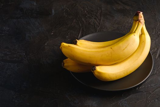 Banana fruits in black plate on dark textured background, angle view copy space