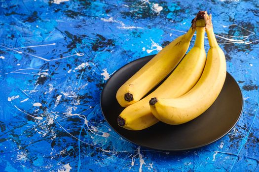 Banana fruits in black plate on blue textured background, angle view copy space
