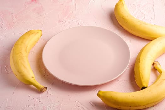 Banana fruits with empty pink plate on pink textured background, angle view copy space