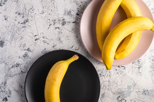 Banana fruits in pink and black plates on white textured background, top view copy space