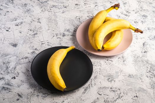 Banana fruits in pink and black plates on white textured background, angle view copy space