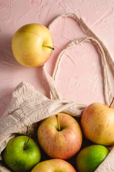 Fresh sweet apples in reusable textile grocery bag on pink textured background, selective focus