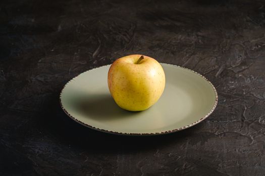 Fresh sweet single apple in grey plate on dark black textured background, angle view