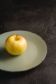 Fresh sweet single apple in grey plate on dark black textured background, angle view