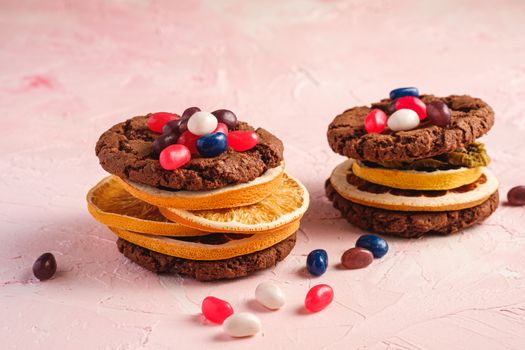 Homemade oat chocolate cookies sandwich with dried citrus fruits and juicy jelly beans on textured pink background, angle view