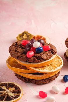 Homemade oat chocolate cookies sandwich with dried citrus fruits and juicy jelly beans on textured pink background, angle view