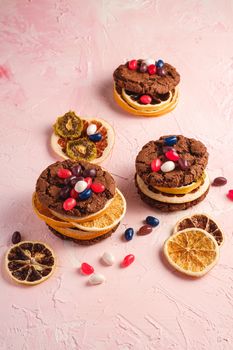 Homemade oat chocolate cookies sandwich with dried citrus fruits and juicy jelly beans on textured pink background, angle view