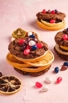 Homemade oat chocolate cookies sandwich with dried citrus fruits and juicy jelly beans on textured pink background, angle view