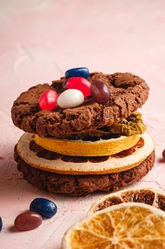 Homemade oat chocolate cookies sandwich with dried citrus fruits and juicy jelly beans on textured pink background, angle view