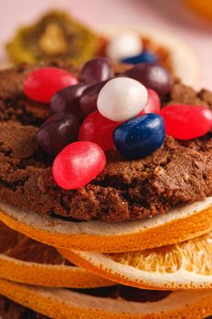 Homemade oat chocolate cookies sandwich with dried citrus fruits and juicy jelly beans on textured pink background, angle view macro