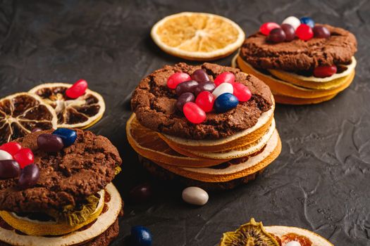 Homemade oat chocolate cookies sandwich with dried citrus fruits and juicy jelly beans on textured dark black background, angle view