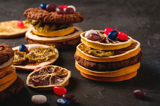 Homemade oat chocolate cookies sandwich with dried citrus fruits and juicy jelly beans on textured dark black background, angle view macro