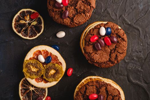 Homemade oat chocolate cookies sandwich with dried citrus fruits and juicy jelly beans on textured dark black background, top view