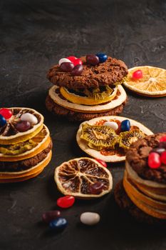 Homemade oat chocolate cookies sandwich with dried citrus fruits and juicy jelly beans on textured dark black background, angle view