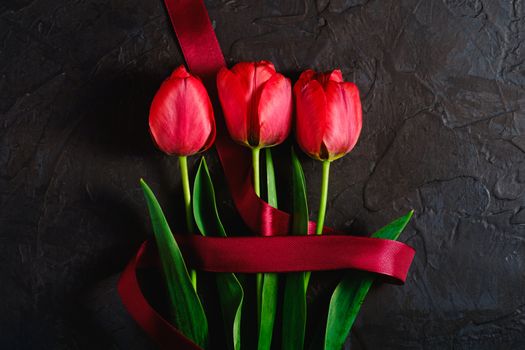 Red bunch of tulip flowers with red ribbon on textured black background, top view copy space
