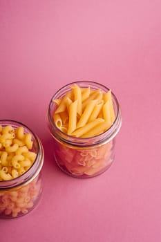 Two glass jars with variety of uncooked golden wheat pasta on minimal pink background, angle view copy space