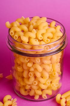 One glass jar with cavatappi uncooked golden wheat curly pasta on minimal pink background, angle view macro