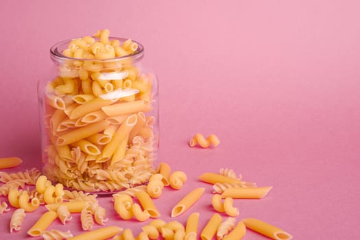 One glass jar with variety of uncooked golden wheat pasta on minimal pink background, angle view macro, copy space