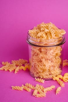 One glass jar with fusilli uncooked golden wheat pasta on minimal pink background, angle view macro