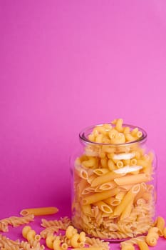 One glass jar with variety of uncooked golden wheat pasta on minimal pink background, angle view macro, copy space