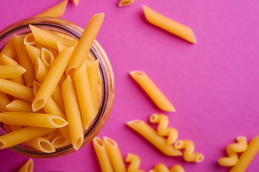 One glass jar with penne uncooked golden wheat tube pasta on minimal pink background, top view macro
