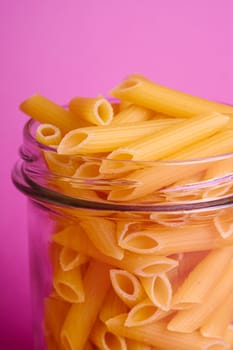 One glass jar with penne uncooked golden wheat tube pasta on minimal pink background, angle view macro