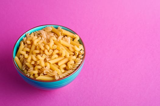Blue bowl with variety of uncooked golden wheat pasta on minimal pink background, angle view copy space
