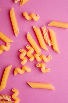 Scattered variety of uncooked golden wheat pasta on minimal pink background, top view