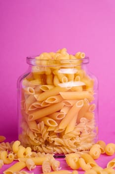 One glass jar with variety of uncooked golden wheat pasta on minimal pink background, angle view macro
