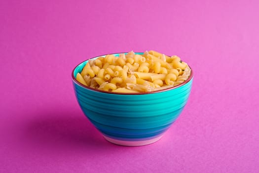 Blue bowl with variety of uncooked golden wheat pasta on minimal pink background, angle view