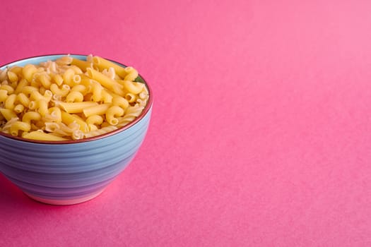 Blue bowl with variety of uncooked golden wheat pasta on minimal pink background, angle view copy space