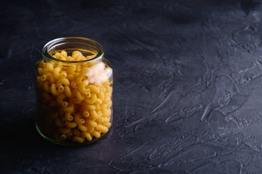 One glass jar with cavatappi uncooked golden wheat curly pasta on textured dark black background, angle view copy space