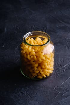 One glass jar with cavatappi uncooked golden wheat curly pasta on textured dark black background, angle view