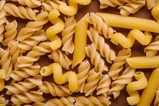 Scattered variety of uncooked golden wheat pasta on minimal brown background, top view