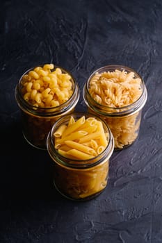 Three glass jars with variety of uncooked golden wheat pasta on dark black textured background, angle view