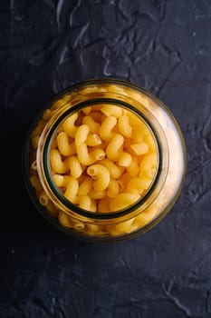 One glass jar with cavatappi uncooked golden wheat curly pasta on textured dark black background, top view