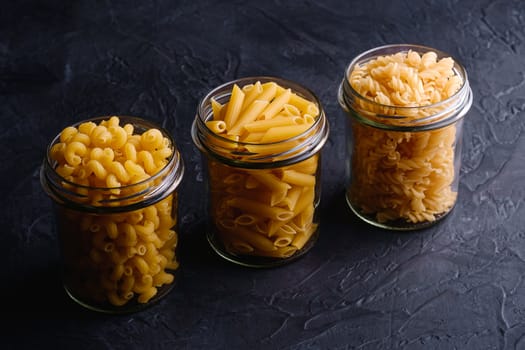Three glass jars in row with variety of uncooked golden wheat pasta on dark black textured background, angle view