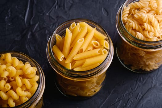 Three glass jars in row with variety of uncooked golden wheat pasta on dark black textured background, angle view macro