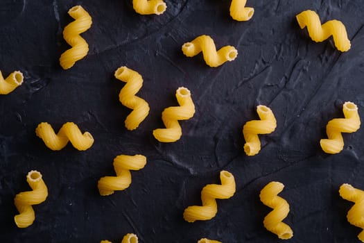 Scattered cavatappi uncooked golden wheat curly pasta pattern on textured dark black background, top view