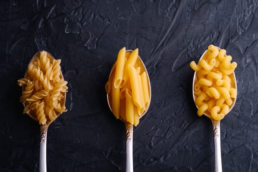 Three cutlery spoons with variety of uncooked golden wheat pasta on dark black textured background, top view