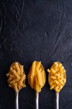 Three cutlery spoons with variety of uncooked golden wheat pasta on dark black textured background, top view copy space