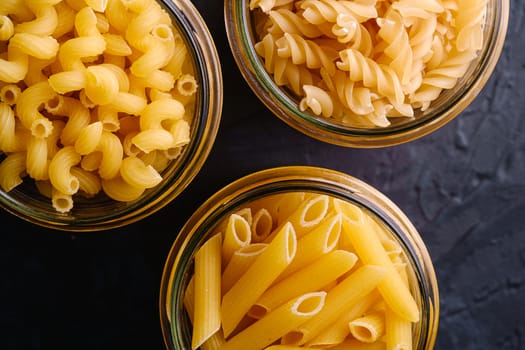 Three glass jars with variety of uncooked golden wheat pasta on dark black textured background, top view macro